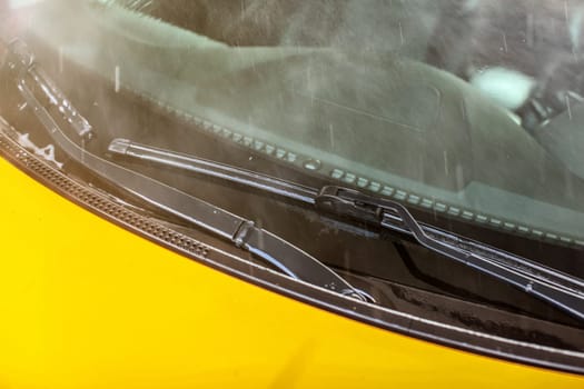 Windshield of yellow car washed in carwash, water flowing over front glass and wiper.