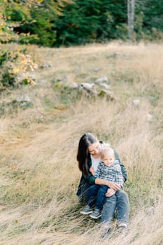 Mom hugs a little girl sitting in her arms on the grass in the park. High quality photo