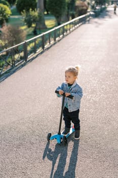 Little girl on a three-wheeled scooter stands on the road in the garden and looks into the distance. High quality photo