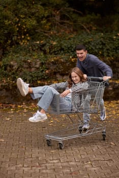 Autumn Joyride: Boy Rolling Girl in Shopping Cart amid Nature's Beauty. playful couple, autumnal adventure, outdoor fun, seasonal joy, laughter and love, romantic play, nature's embrace, joyful moments, couple goals, autumn magic.