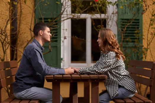 Happy smiling young dating couple having coffee together and enjoying life sitting at table holding hands in street cafe on summer day. Pretty man and woman spending time together enjoying weekend. Dating, young fashion elegant stylish couple sitting at a table in a cafe , gently hold hands, on streets of old european city. American beautiful man hugging caucasian woman and sitting at cafe, hat on table. Concept of happy international couple and dating.