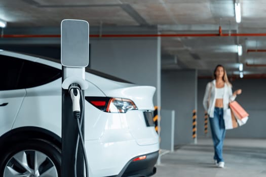 Young woman travel with EV electric car to shopping center parking lot charging in downtown city showing urban sustainability lifestyle by green clean rechargeable energy of electric vehicle innards