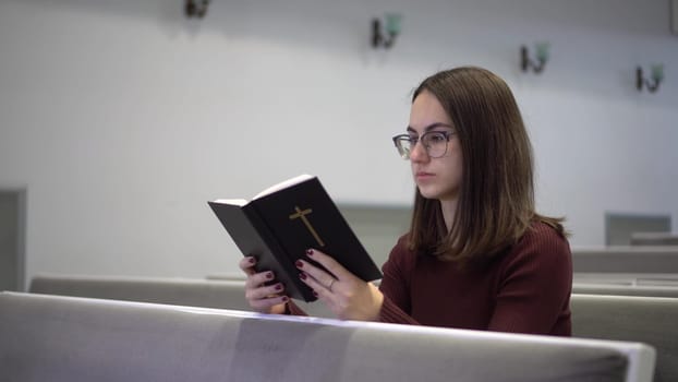 A young woman reads the Bible while sitting on a church bench. A Protestant girl with glasses reads the Bible in church. 4k
