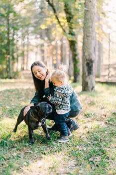Little girl hugging laughing mom stroking dog in park. High quality photo