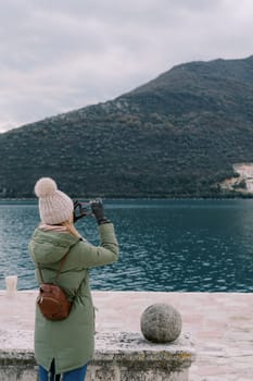 Girl stands by the stone fence on the seashore and takes pictures of the mountains with a smartphone. Back view. High quality photo