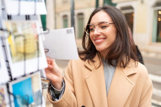 Smiling woman wear coat and glasses looks at postcards on street. Female traveler choosing postcard standing outdoor in european city at autumn.