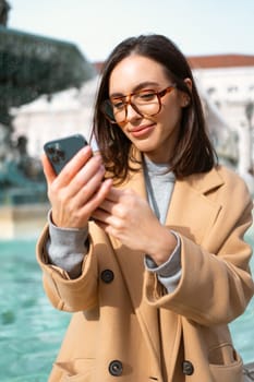 Woman wear glasses looks into smartphone against background of fountain. Female on European city street in coat stands near fountain in autumn looking at phone. Vertical photo