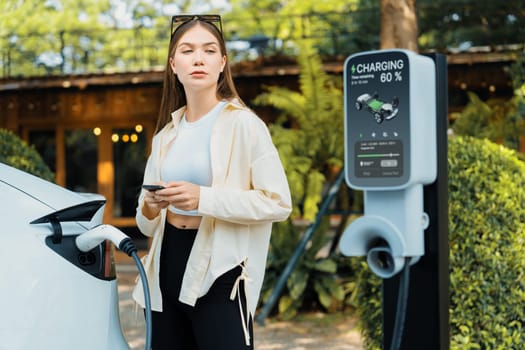 Sustainable urban commute with young woman pay electricity for EV electric car recharging at outdoor cafe in springtime garden, green city sustainability and environmental friendly EV car. Expedient