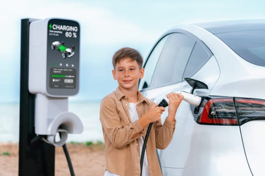 Little boy recharging eco-friendly electric car from EV charging station. EV car road trip travel by the seashore by alternative vehicle powered by clean renewable and sustainable energy. Perpetual