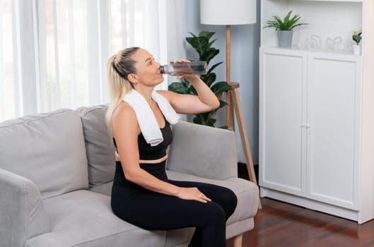 Athletic and sporty senior woman taking a break and sitting on sofa while drinking water after home exercise as concept of healthy fit body lifestyle after retirement. Clout