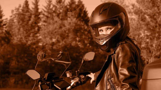 Biker girl riding touring motorbike on forest road. Image toned in Peach Fuzz color of the year 2024. Beautiful woman with red hair in leather jacket sitting on motorcycle in the park