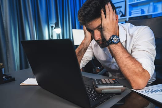 Stressful businessman with headache analyzing paperwork project at night time, waiting email send back for startup product. Concept of overworked design on neon blue light modern office. Sellable.