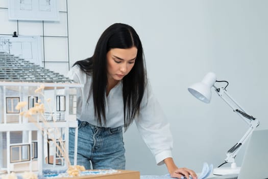 Portrait of Young beautiful caucasian architect focus on comparing house structure between house model and blueprint and laptop placed on meeting table. Check, inspect, estimate concept. Immaculate.