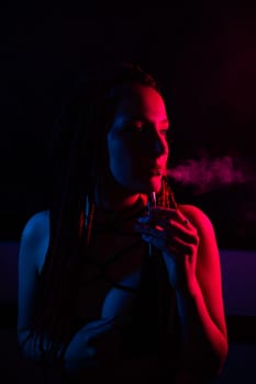 Profile of caucasian girl with dreadlocks smokes a vape in red blue light