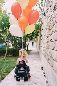 Little girl is driving a toy car with tied balloons around the yard. Back view. High quality photo