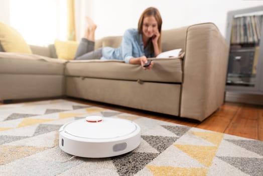 Robotic vacuum cleaner cleaning the room while woman relaxing on sofa. Woman controlling vacuum with remote control.