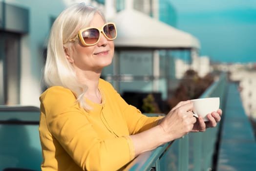 Cheerful, elderly woman enjoying sunny day on balcony. Senior people activity on retirement concept