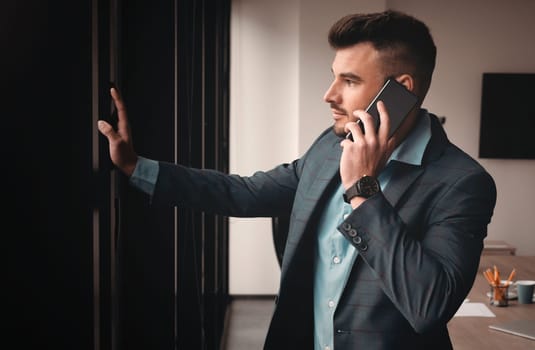 Portrait of a businessman in the office. Handsome man is standing by the window with phone
