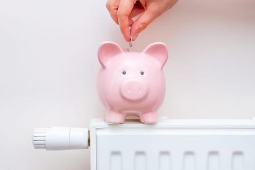 Radiator at home, the concept of rising heating prices. Hand puts the coin into the piggy bank