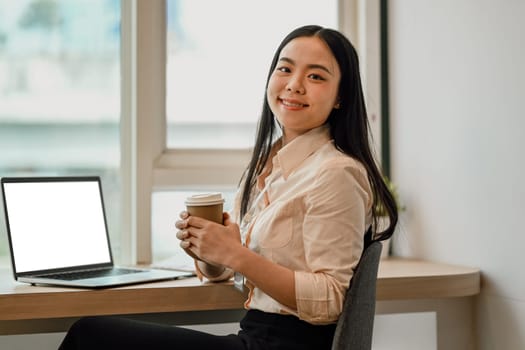 Portrait of cheerful Asian female office worker sitting at workplace and smiling to camera.