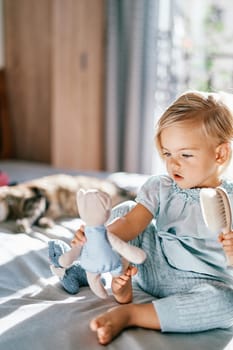 Little girl holds a teddy bear in her outstretched hand with a comb in her other hand while sitting on the bed. High quality photo