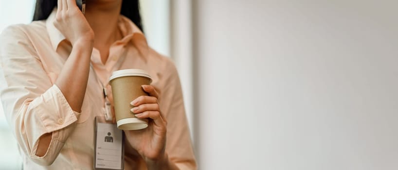Young woman employee holding a paper cup of hot coffee and talking on mobile phone.