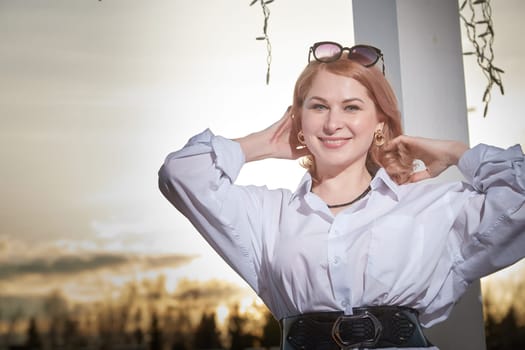 Portrait of Beautiful girl with red hair in white shirt in open wooden pavillion in village or small town. Young slender woman and sky with clouds on background on autumn, spring or summer evening