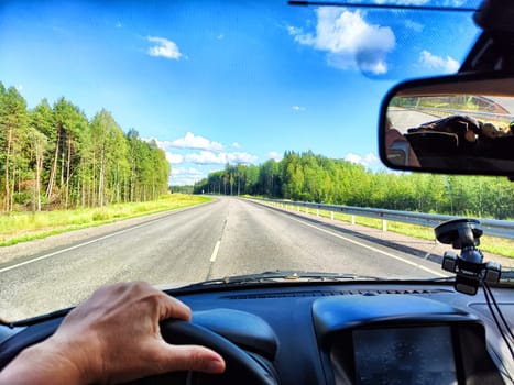 Car salon, steering wheel, hand of woman, view on nature landscape. View from seat of driver on Road, forest, blue sky, white clouds at sunny day. Concept of single trip of female traveller