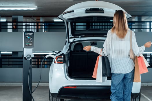 Young woman travel with EV electric car to shopping center parking lot charging in downtown city showing urban sustainability lifestyle by green clean rechargeable energy of electric vehicle innards