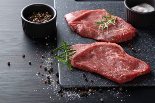 Two fresh raw marbled beef steaks on a cutting board on a wooden background with salt, pepper and rosemary in a rustic style.