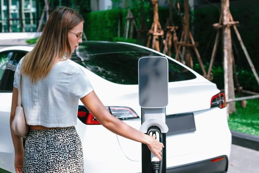 Young woman travel with EV electric car charging in green sustainable city outdoor garden in summer. Urban sustainability lifestyle by green clean rechargeable energy of electric BEV vehicle innards