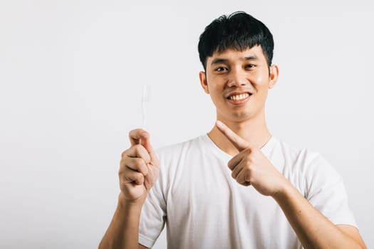 Smiling Asian young man, in a dental health concept, brushes teeth with a toothbrush and points to it with enthusiasm. Studio shot isolated on white background, showing a positive attitude.