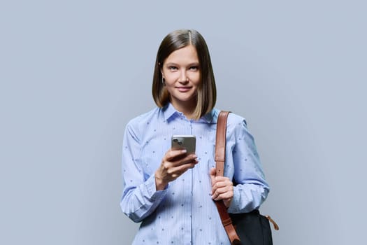 Young woman student with smartphone backpack on grey studio background. Smiling attractive female looking at camera. Using mobile applications apps for work business study leisure, technology, people