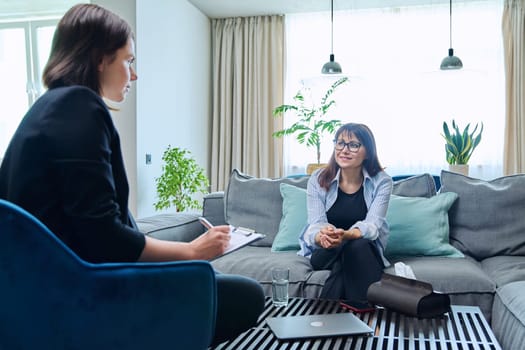 Female psychologist is having therapy session with middle-aged woman patient sitting on sofa in office. Psychology, psychotherapy, therapy, counseling, treatment, mental health concept