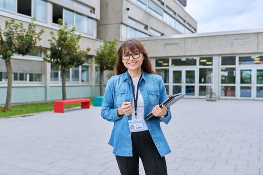 Middle-aged confident woman school teacher, mentor, pedagogue, psychologist, counselor, social worker with digital tablet in hands posing near school building, outdoor