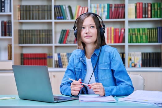 Portrait of smiling young female university student wearing headphones sitting at desk with laptop in library classroom of educational building. Knowledge, education, youth concept