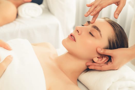 Caucasian couple enjoying relaxing anti-stress head massage and pampering facial beauty skin recreation leisure in dayspa modern light ambient at luxury resort or hotel spa salon. Quiescent