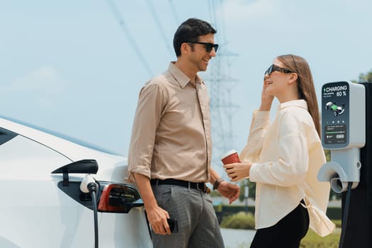 Young couple with coffee recharge EV car battery at charging station connected to electrical industrial power grid tower. Couple with shopping and travel using eco electric car lifestyle. Expedient