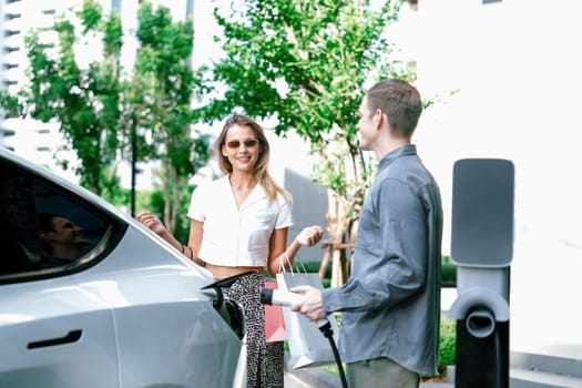 Young couple travel with EV electric car charging in green sustainable city outdoor garden in summer shows urban sustainability lifestyle by green clean rechargeable energy of electric vehicle innards