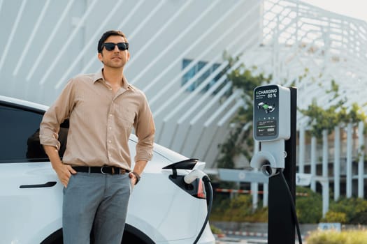 Young man put EV charger to recharge electric car's battery from charging station in city commercial parking lot. Rechargeable EV car for sustainable environmental friendly urban travel. Expedient