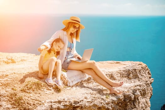 Freelance woman with her daughter working on a laptop by the sea, typing on the keyboard, enjoying the beautiful view, highlighting the idea of remote work