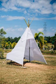 Amidst a lush field a summer wedding tipi offers a charming view. The traditional wigwam adorned with dreamcatcher decorations symbolizes joy and celebration in a natural garden setting.