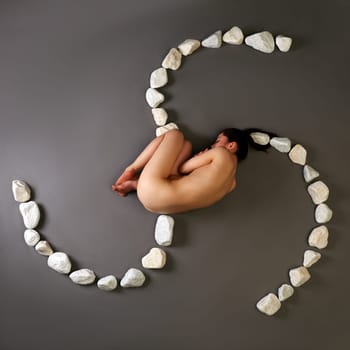 Top view of naked slim woman curled up with stones, on gray background