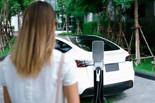 Young woman travel with EV electric car charging in green sustainable city outdoor garden in summer. Urban sustainability lifestyle by green clean rechargeable energy of electric BEV vehicle innards