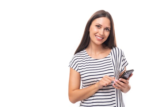 portrait of a young cute casual european woman with long dark hair dressed in a striped t-shirt typing a message on a mobile phone on a white background with copy space.