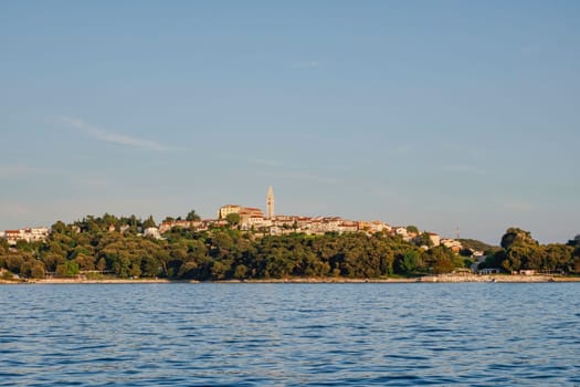 the port town of Vrsar in Croatia seen from a boat from C with the sun on the houses and the church protruding above the town in the middle of the greenery in the woods