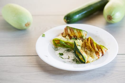 Grilled zucchini pieces with garlic sauce in a white plate. Light gray wooden background.