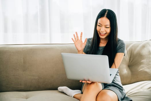 Attractive woman on sofa waving during laptop video call. Smiling teacher makes hello gesture welcoming clients. Engaged in distant meeting fostering connections.