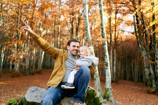 Smiling dad with a little girl on his knees sits on a stump and points his finger at a tree. High quality photo