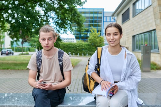 Two high school students guy and girl outdoor, smiling looking at camera, school building background. Education, youth, adolescence concept